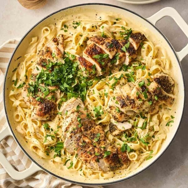 A close up of a large white pan filled with Chicken Alfredo, a dish with long tagliatelle pasta, creamy sauce and sliced herby chicken breast on top, with parsley garnished.