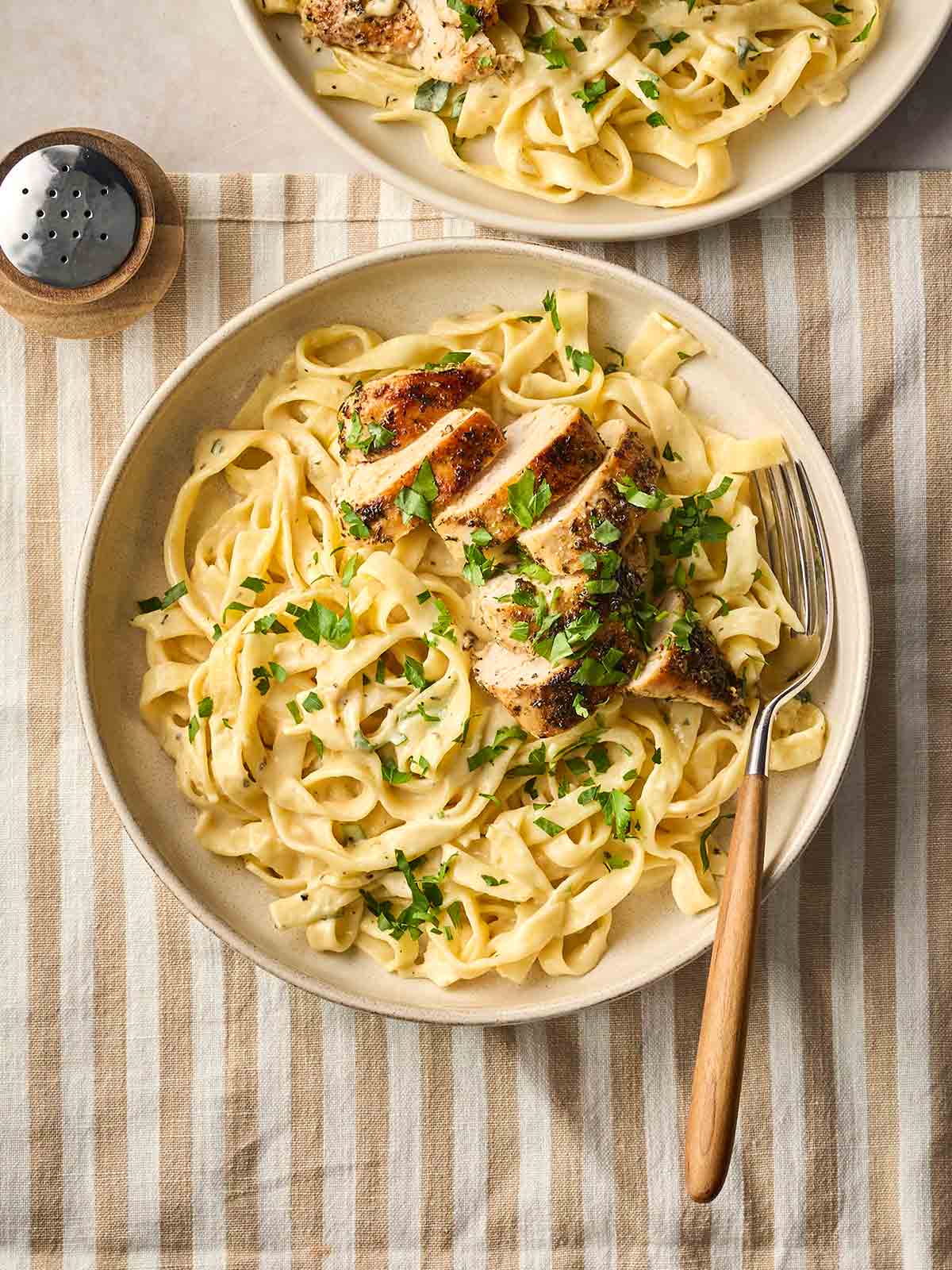 A bowl of Chicken Alfredo on a table with a fork, with another bowl to the top of the image, with a stripy placement underneath and pepper to the side.