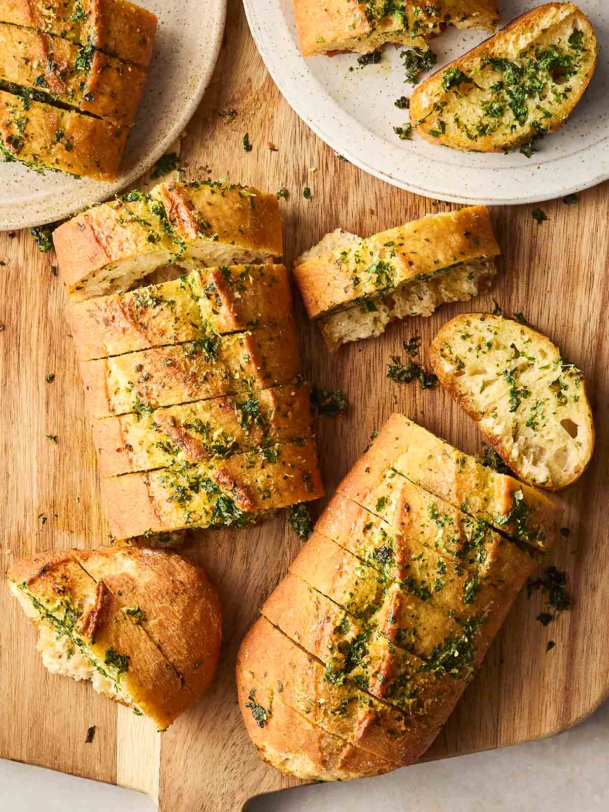 A bread board filled with sliced homemade garlic bread, with two plates with a couple of pieces on.