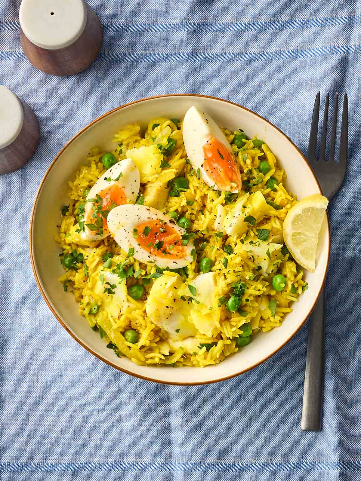 A place setting with a blue cloth, with a bowl of Kedgeree and a fork to the side and salt and pepper above.