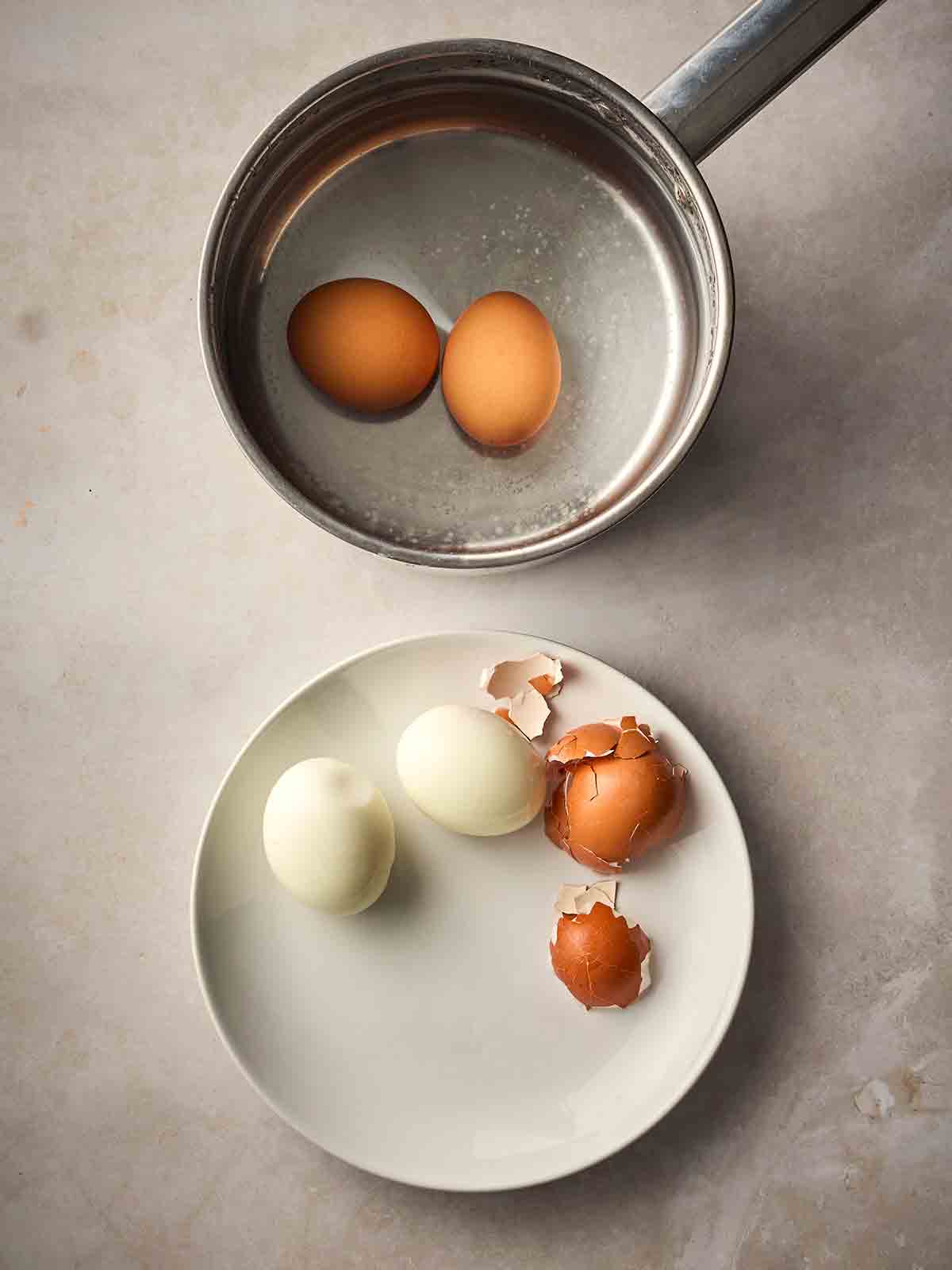 Two boiled eggs in a pan with a plate to side of peeled boiled eggs.