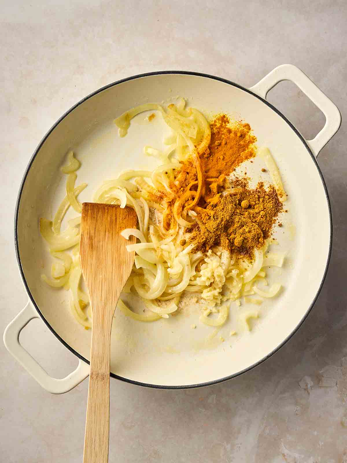A white pan filled with sliced onions and spices, ready to be mixed together with a wooden spatula.