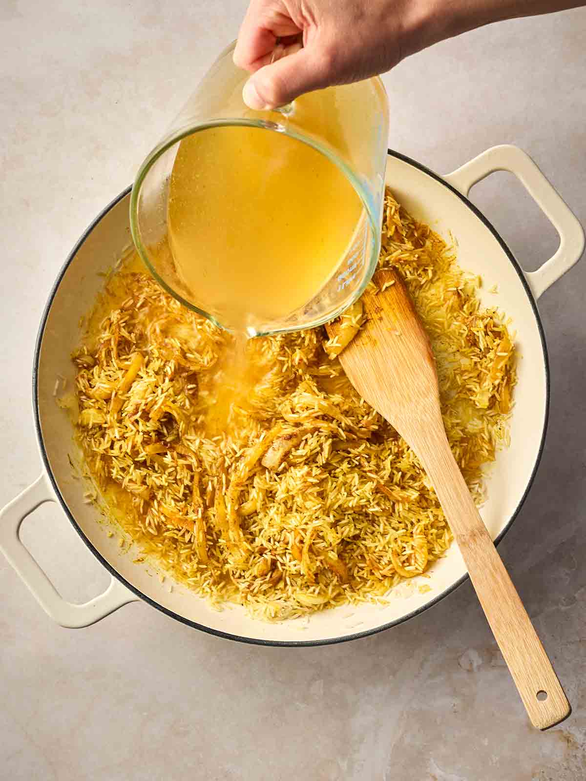 A hand pouring stock into a dish with rice, spices and onion.