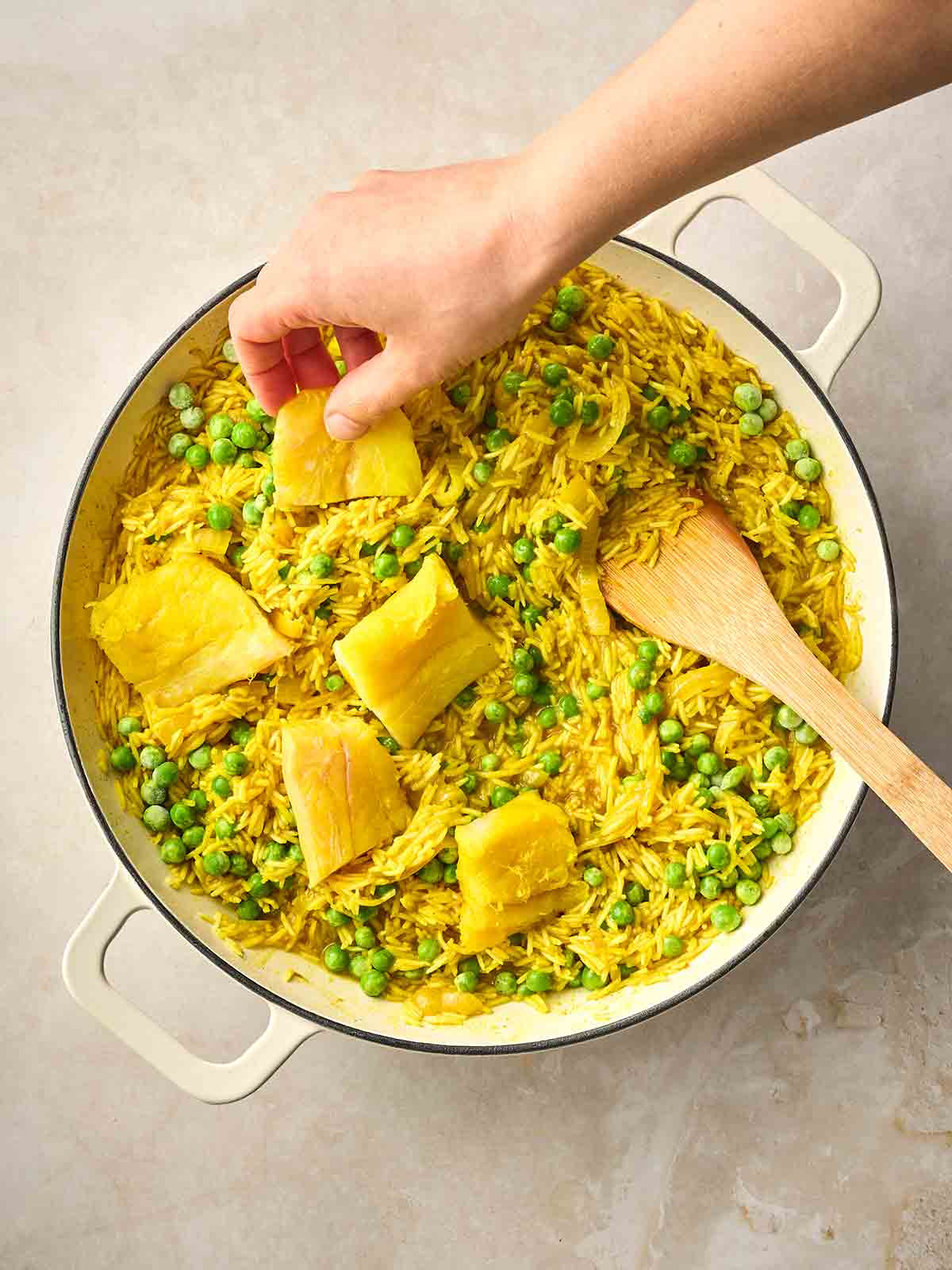 A hand placing pieces of smoked haddock with peas into a pan with curried rice for a Kedgeree dish.