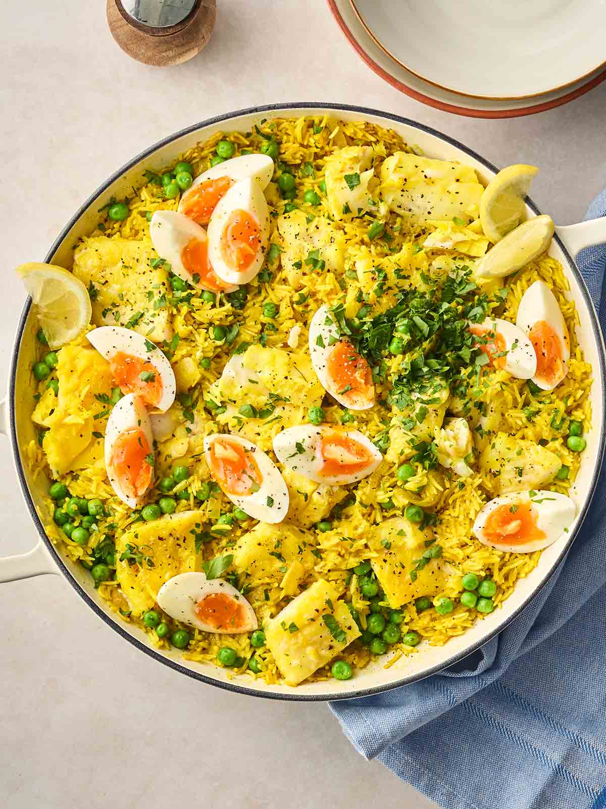 A large pan of Kedgeree with fish, rice and boiled eggs, garnished with lemon wedges and parsley, on a table, ready to serve.