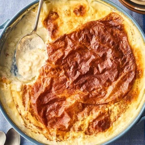 A traditional baked rice pudding in a pan with a golden skin on top, ready to be served with a spoon into bowls. With a blue table cloth in the background.