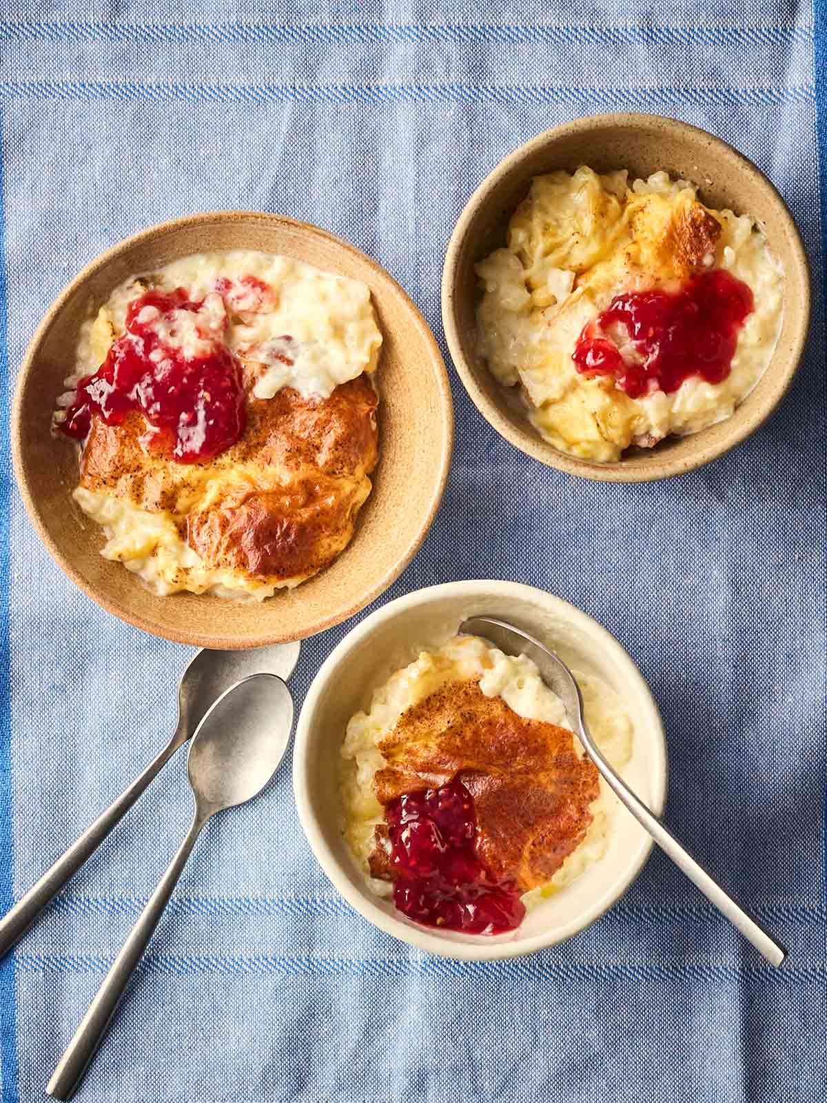 Three bowls of traditional vanilla rice pudding with spoons and jam on top, all on a blue tablecloth.