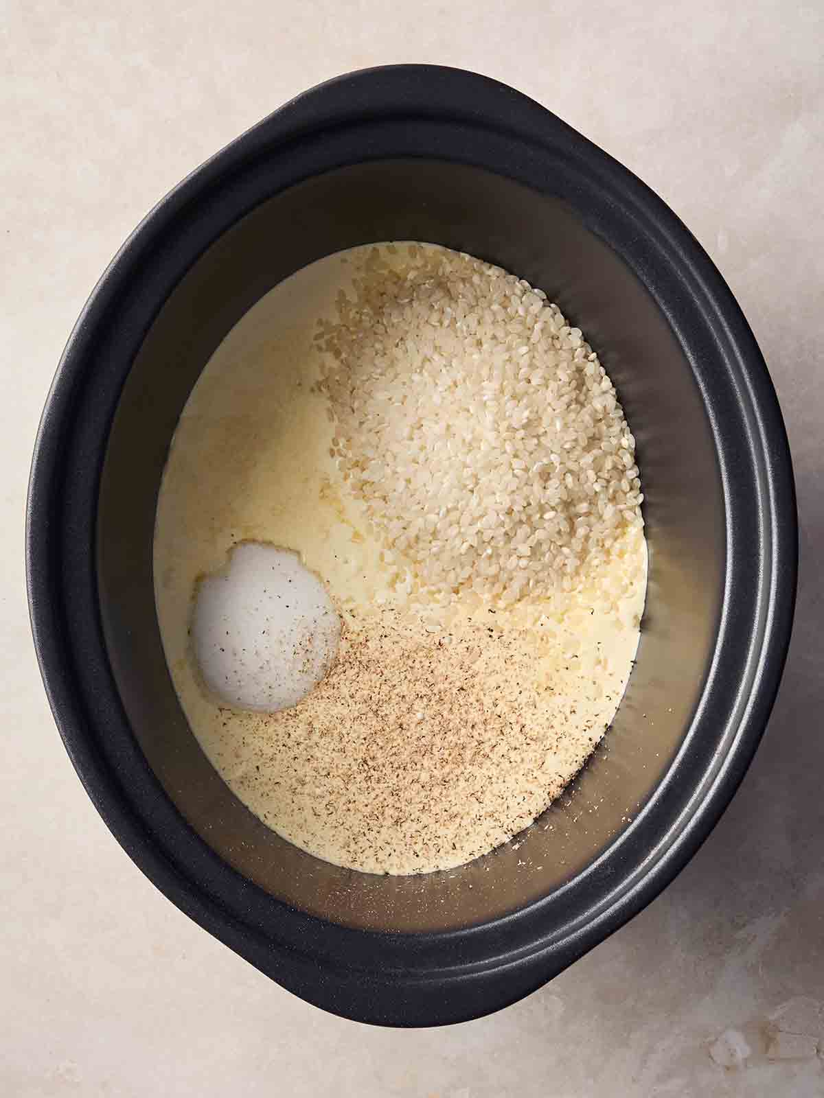 A slow cooker pan on a white surface with uncooked ingredients for rice pudding inside.