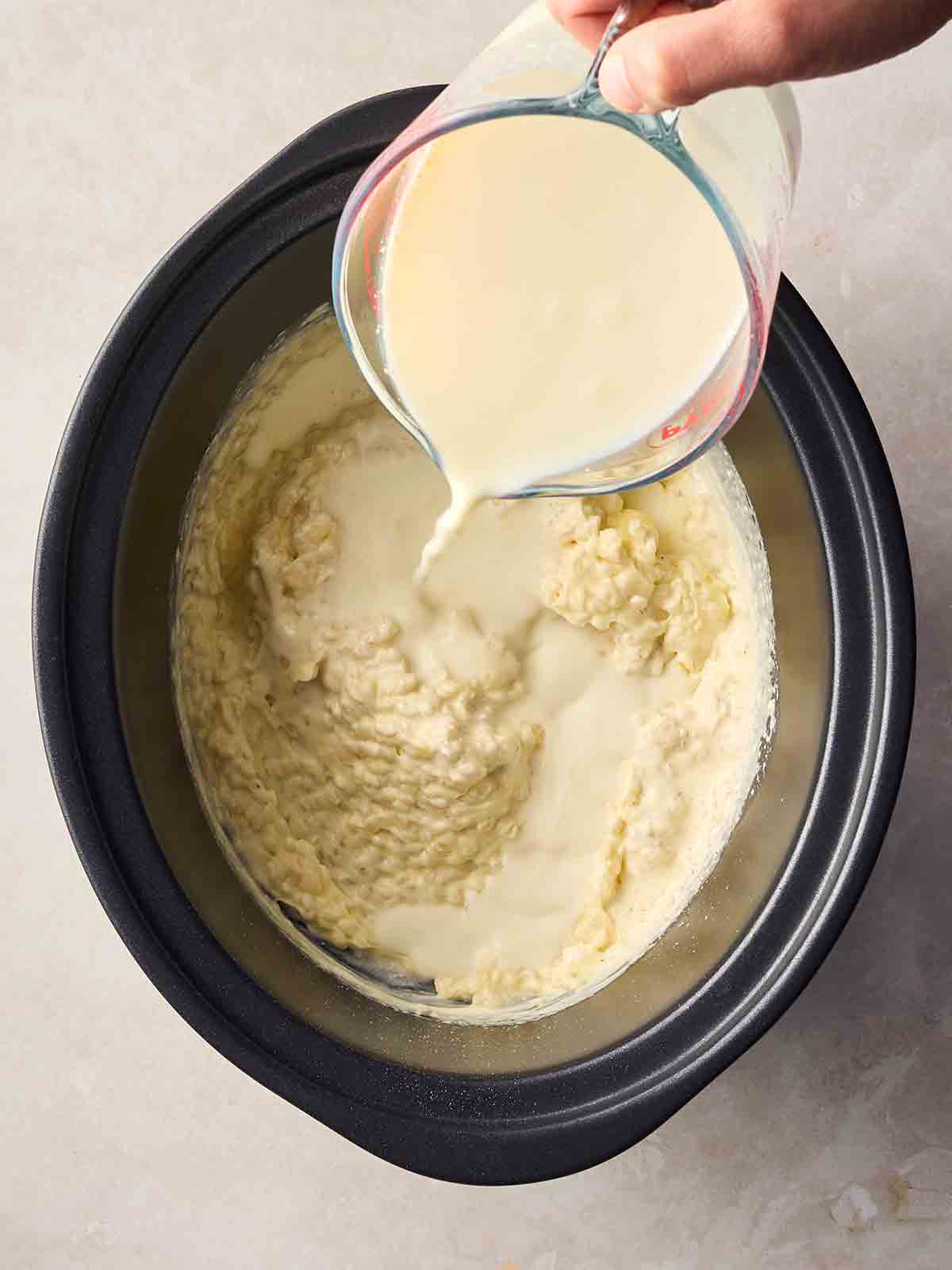 A crock pot pan with cooked rice pudding inside, with a hand pouring more cream into it.