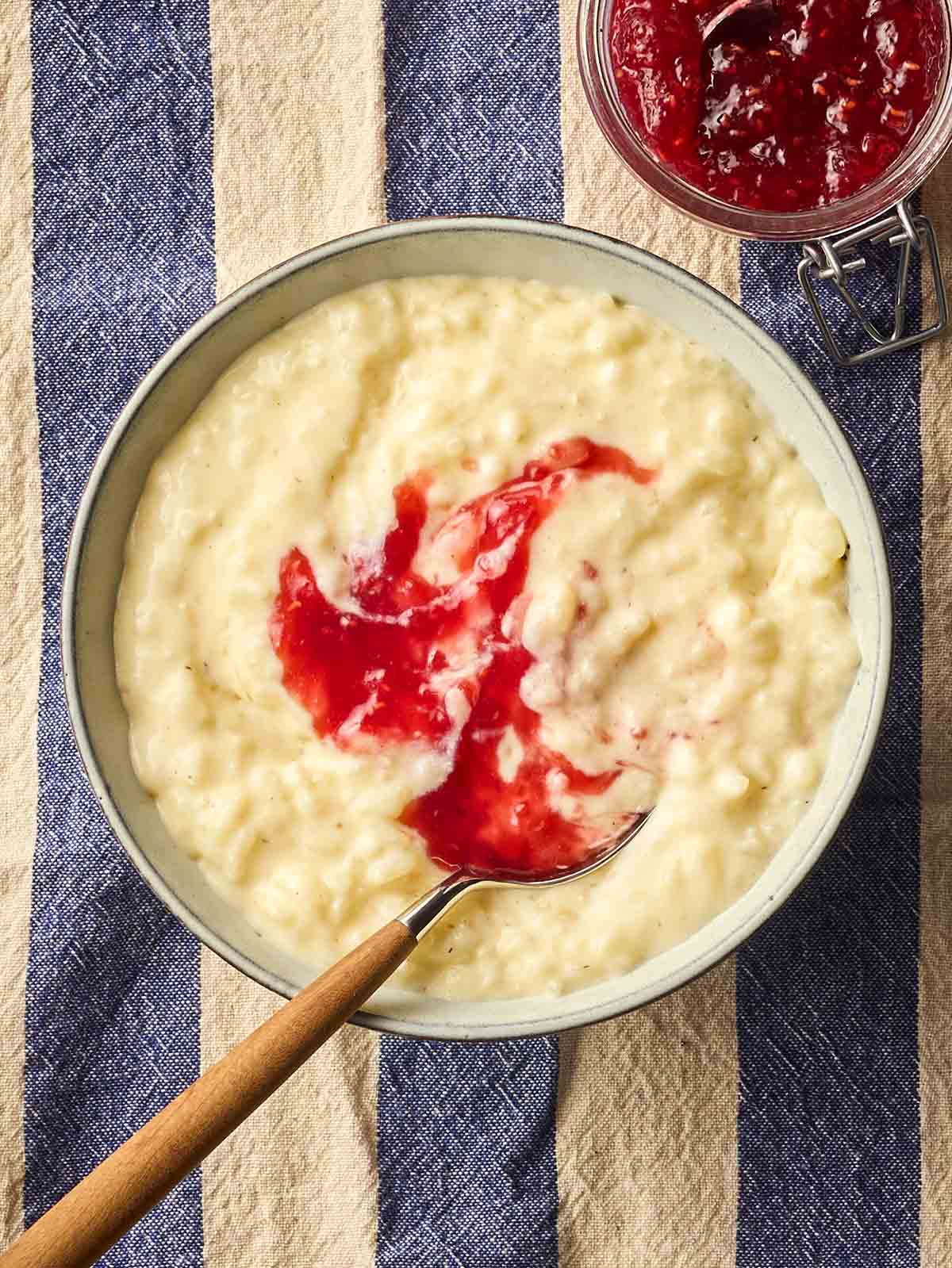 A bowl of homemade slow cooker rice pudding, with jam on top with a spoon, on a white and blue stripy tablecloth.