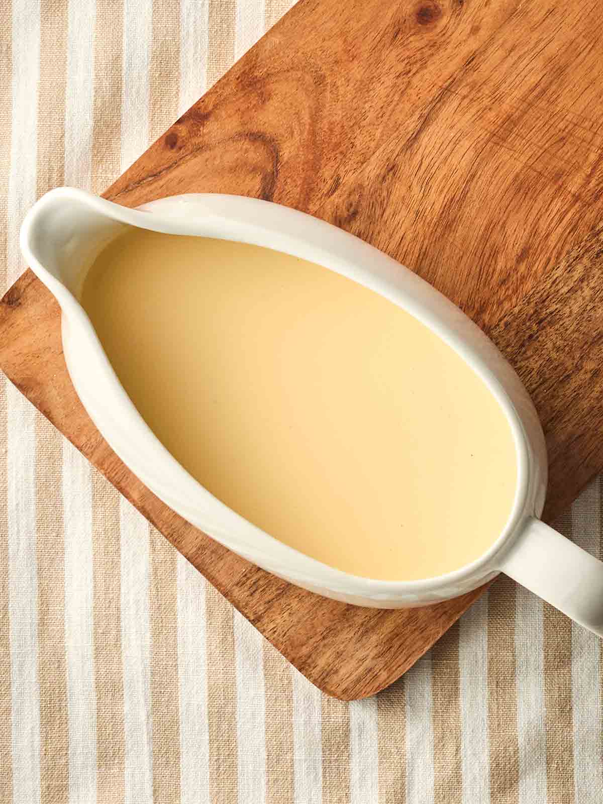 A jug of homemade custard on a board, on a white and beige stripy tablecloth.