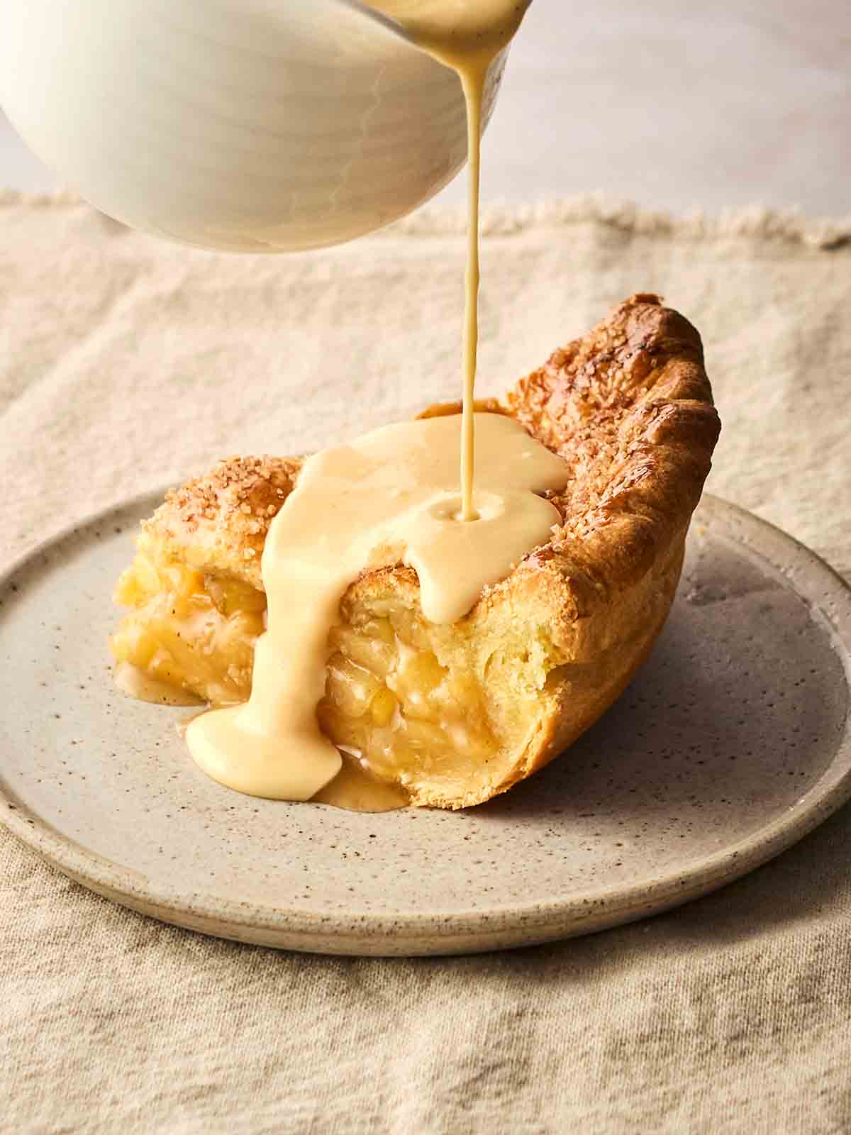 A slice of apple pie on a plate, with custard pouring onto it from a jug above.