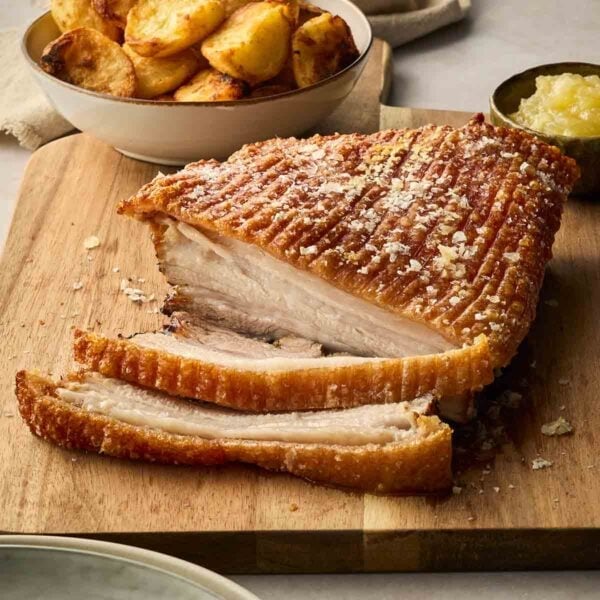 A cooked joint of pork belly with crackling on a wooden serving board, with a small pot of apple sauce and a bowl of roast potatoes.