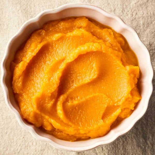 A close up of bright orange pumpkin puree in a white scalloped-edged bowl, on a linen cloth.