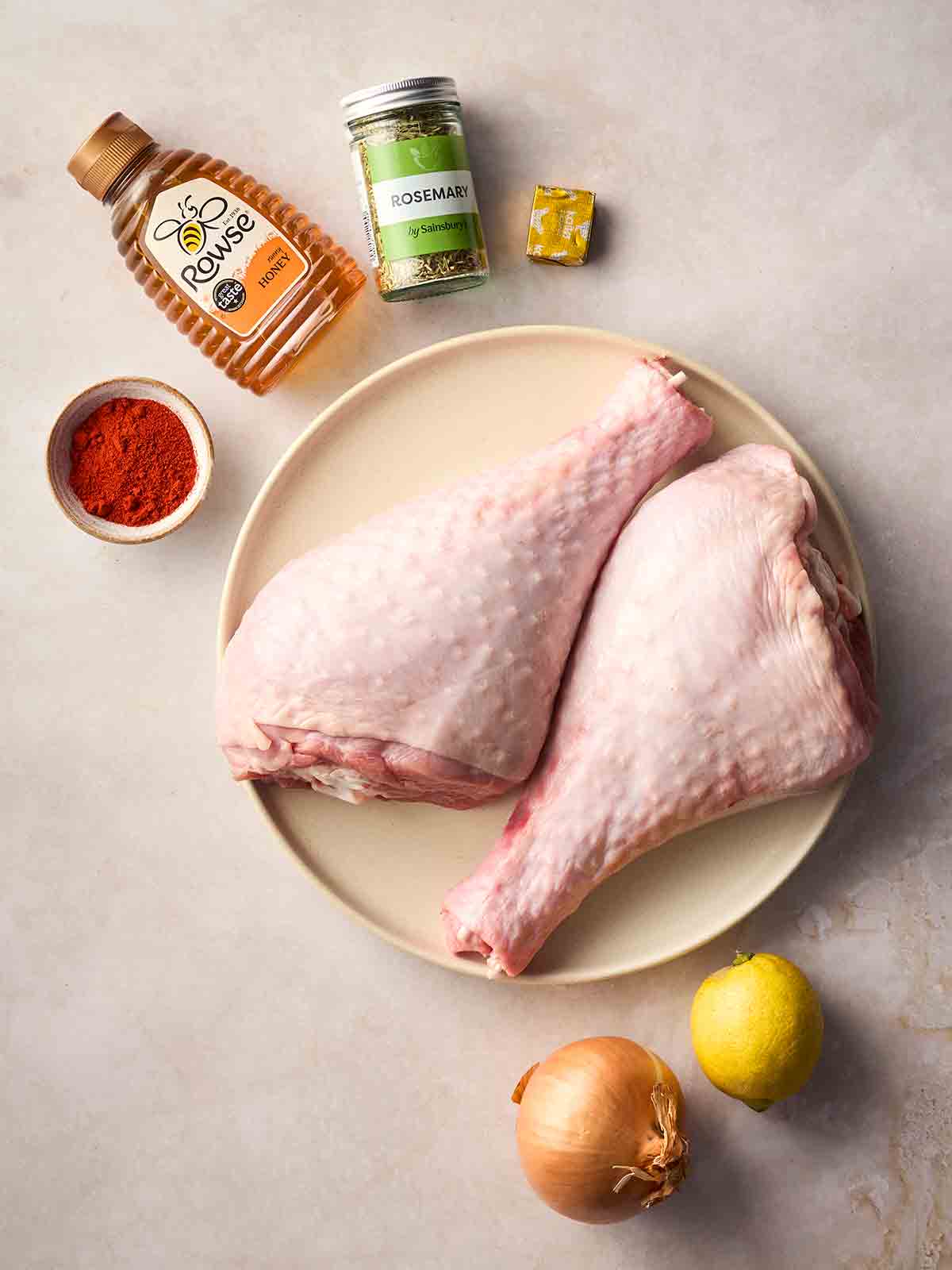 Two raw turkey legs on a plate on a white surface, surrounded by other ingredients to roast them.
