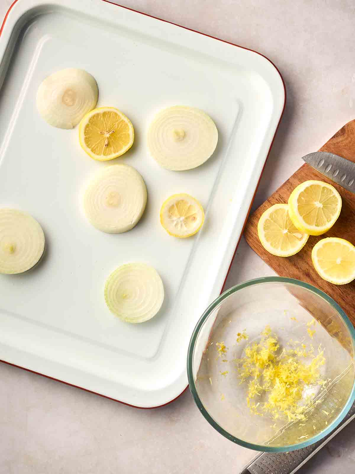 A roasting tray with slices of onion and lemon, with lemon zest in a glass bowl to the side, and a board with a knife and lemons on.