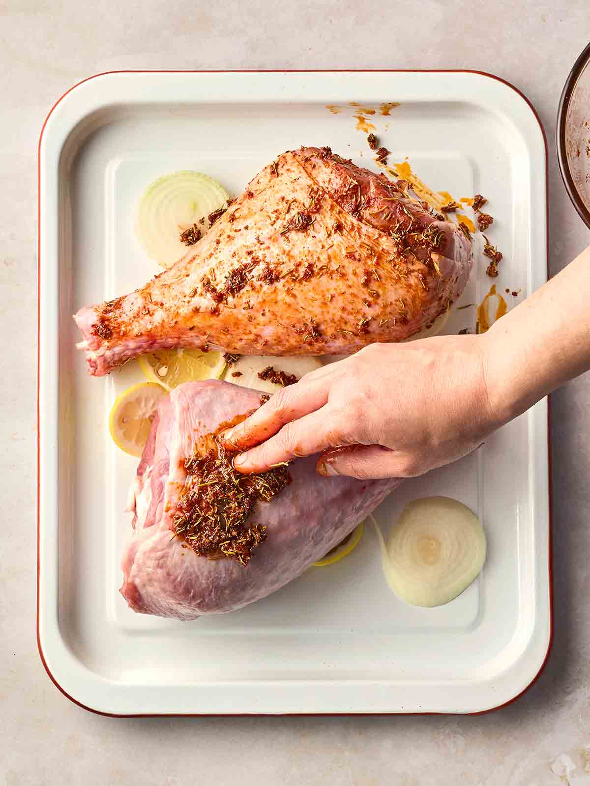 A hand rubbing a marinade onto turkey legs on a roasting tray.