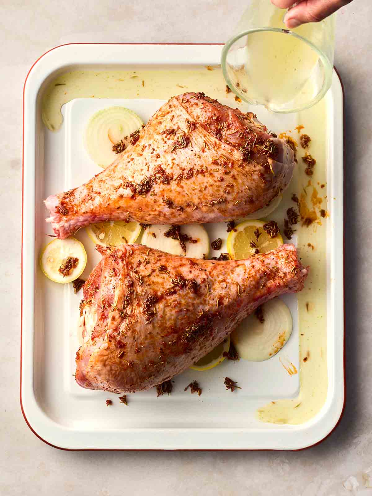 A hand pouring lemon juice from a glass jug over the top of two raw turkey legs, ready to roast.
