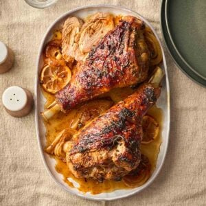 A platter with two marinated roasted turkey legs on, surrounded by a sauce with lemon and onion.
