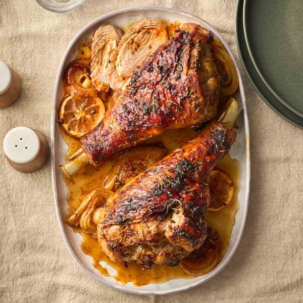 A platter with two marinated roasted turkey legs on, surrounded by a sauce with lemon and onion.