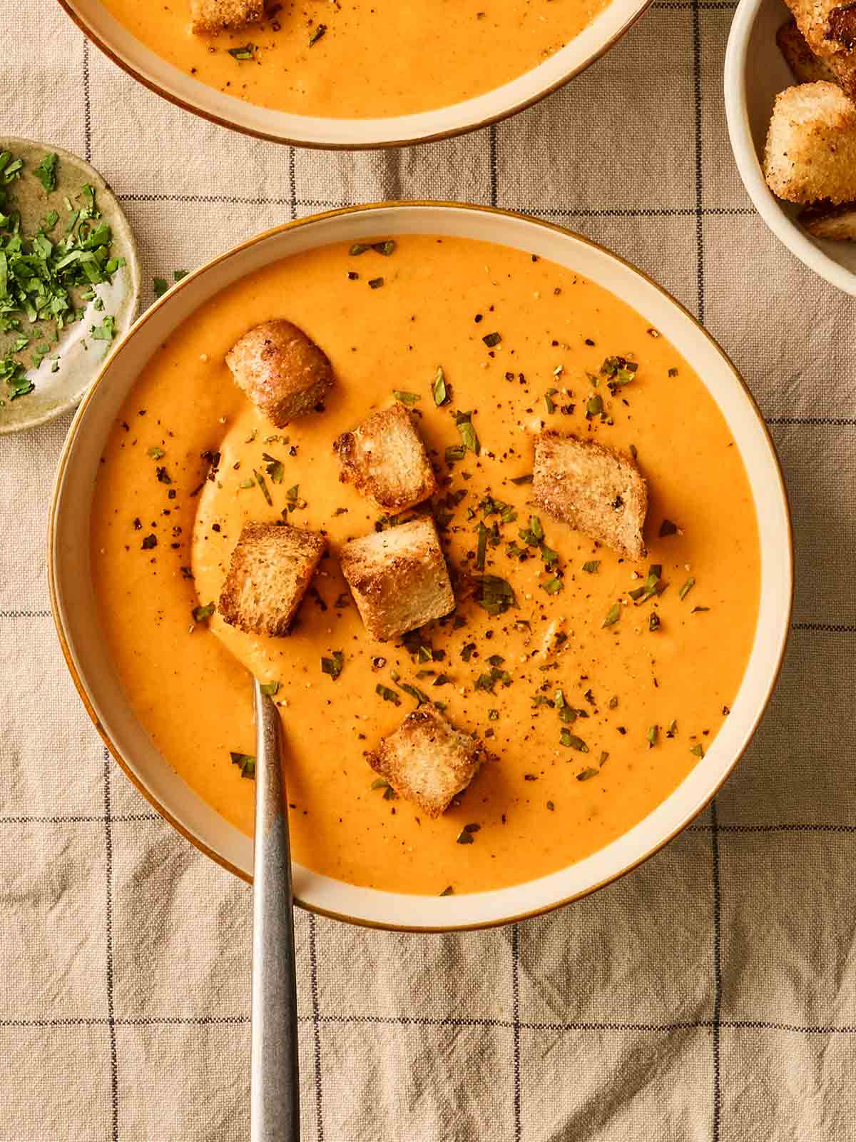 A bowl of bright orange sweet potato soup with croutons on top, on a table cloth that is patterned with squares.